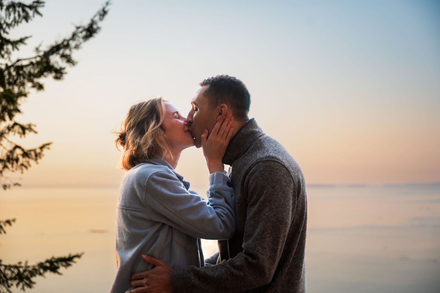 A couple embracing and kissing against a sunset backdrop over calm waters.