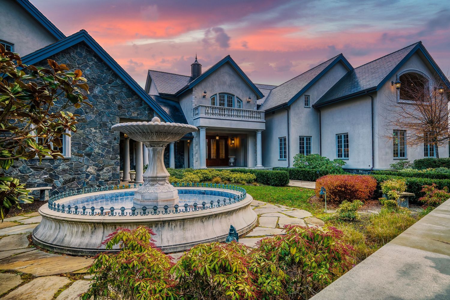 Luxurious estate entrance featuring ornate fountain, stone facade and manicured landscaping at sunset.