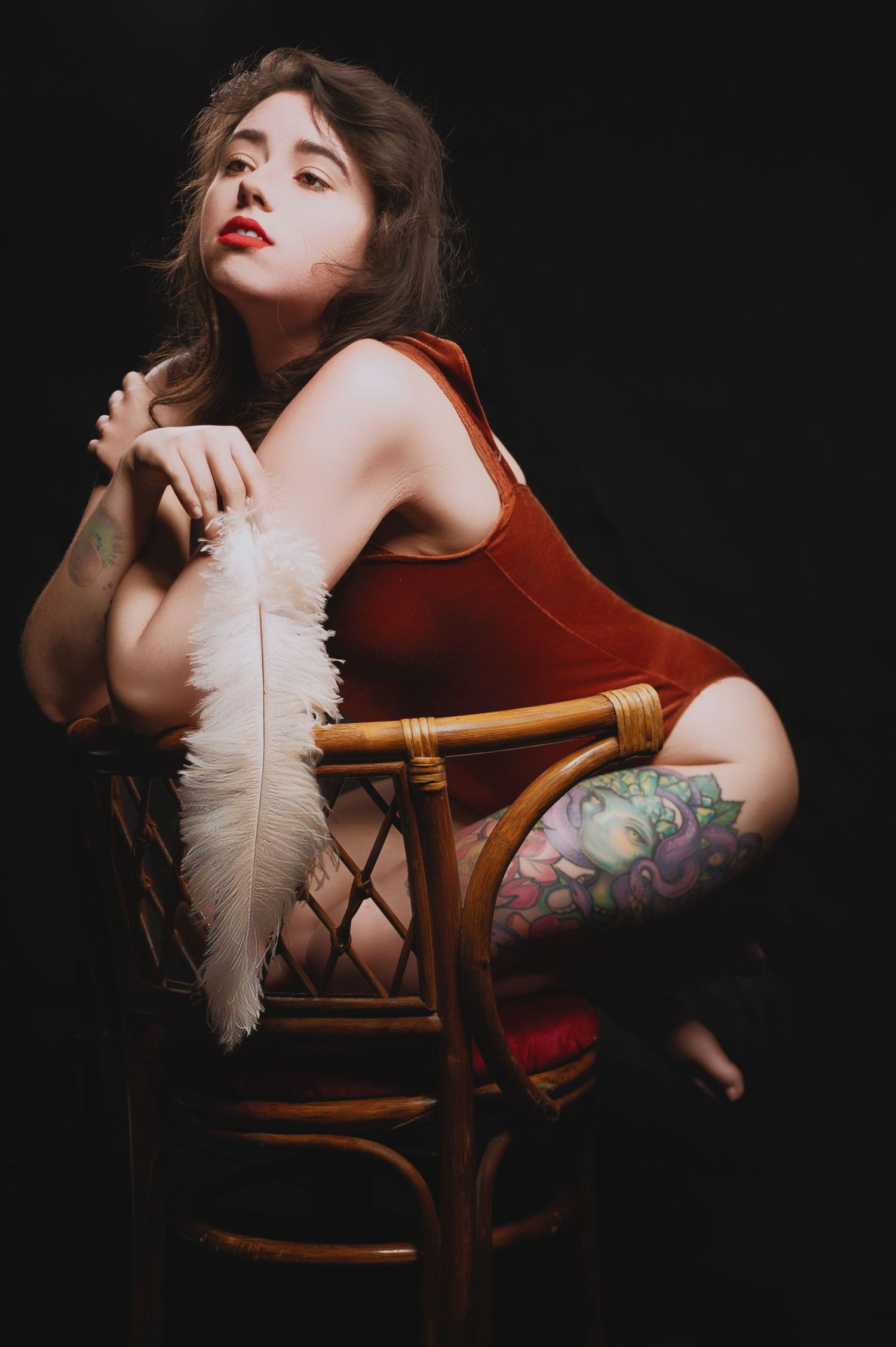 Artistic boudoir portrait with white feather and wooden chair against dark background.