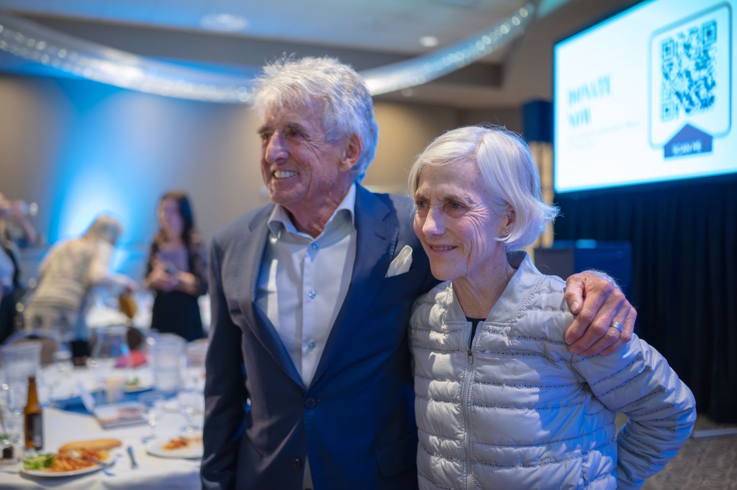 Two people posing together at a formal dinner event with projection screen visible in background.