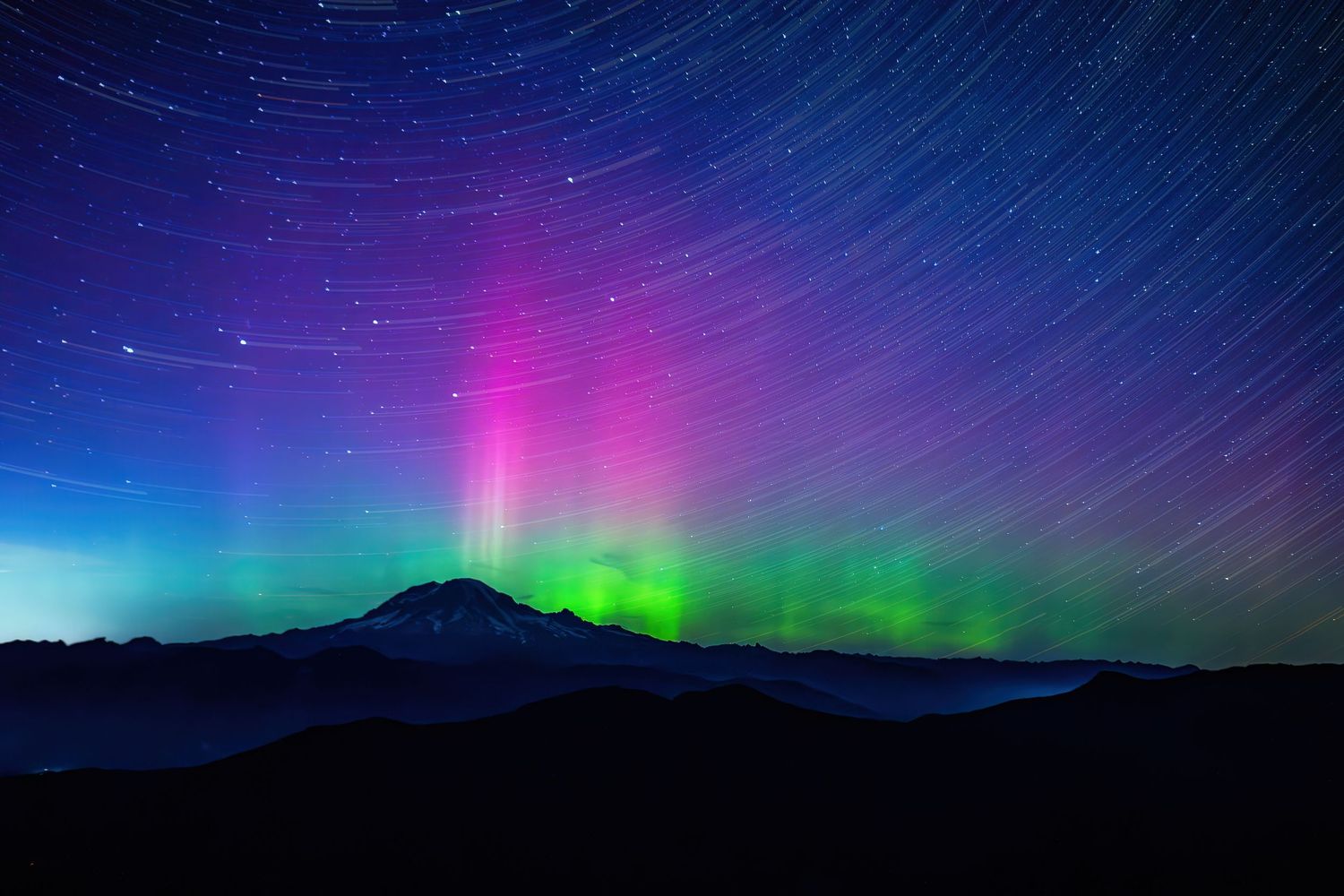Vibrant aurora borealis with pink and green hues illuminates night sky above dark mountain silhouette with star trails.