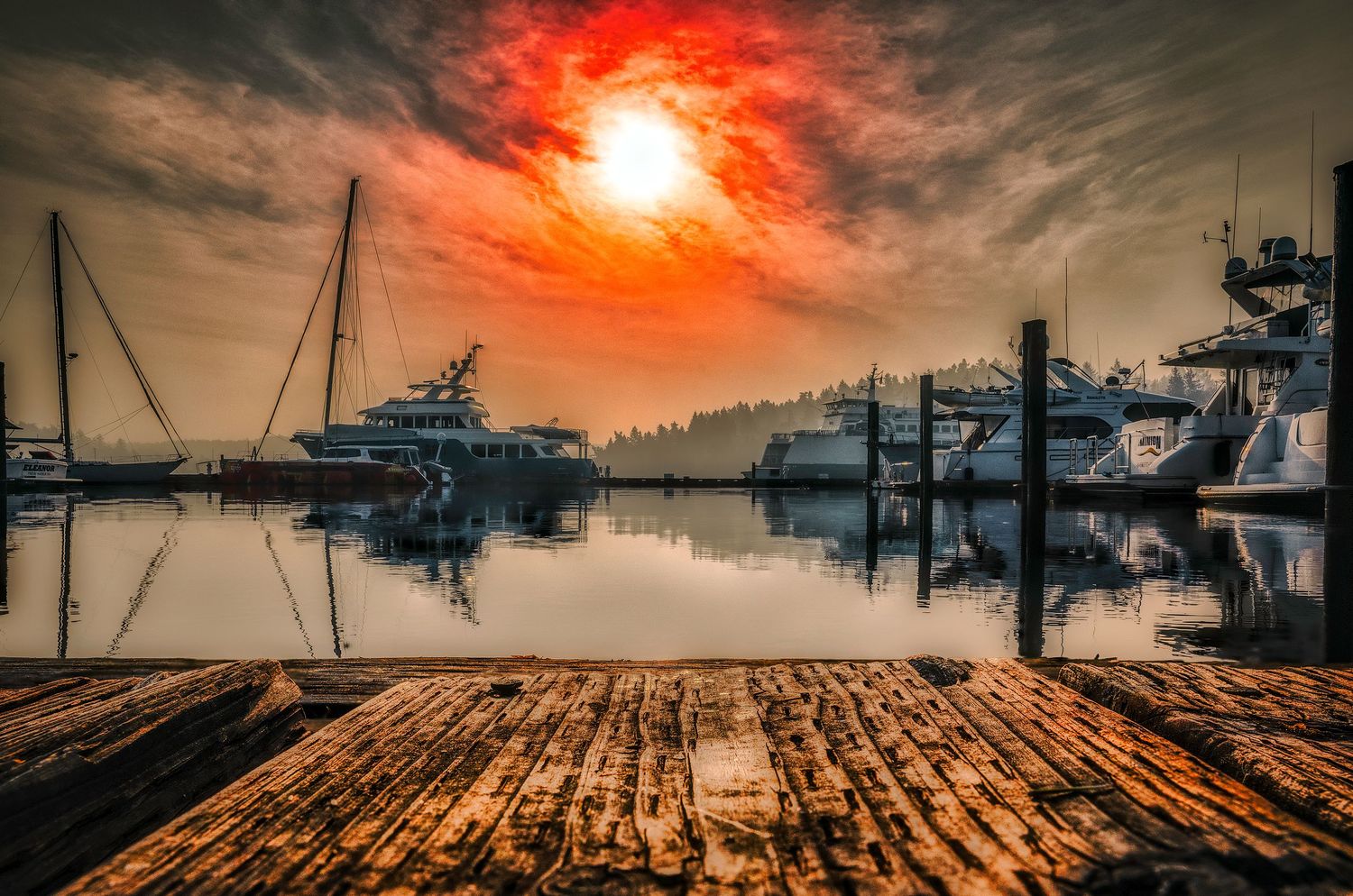 Marina dock with boats at sunset featuring dramatic red sky reflected in calm harbor waters.