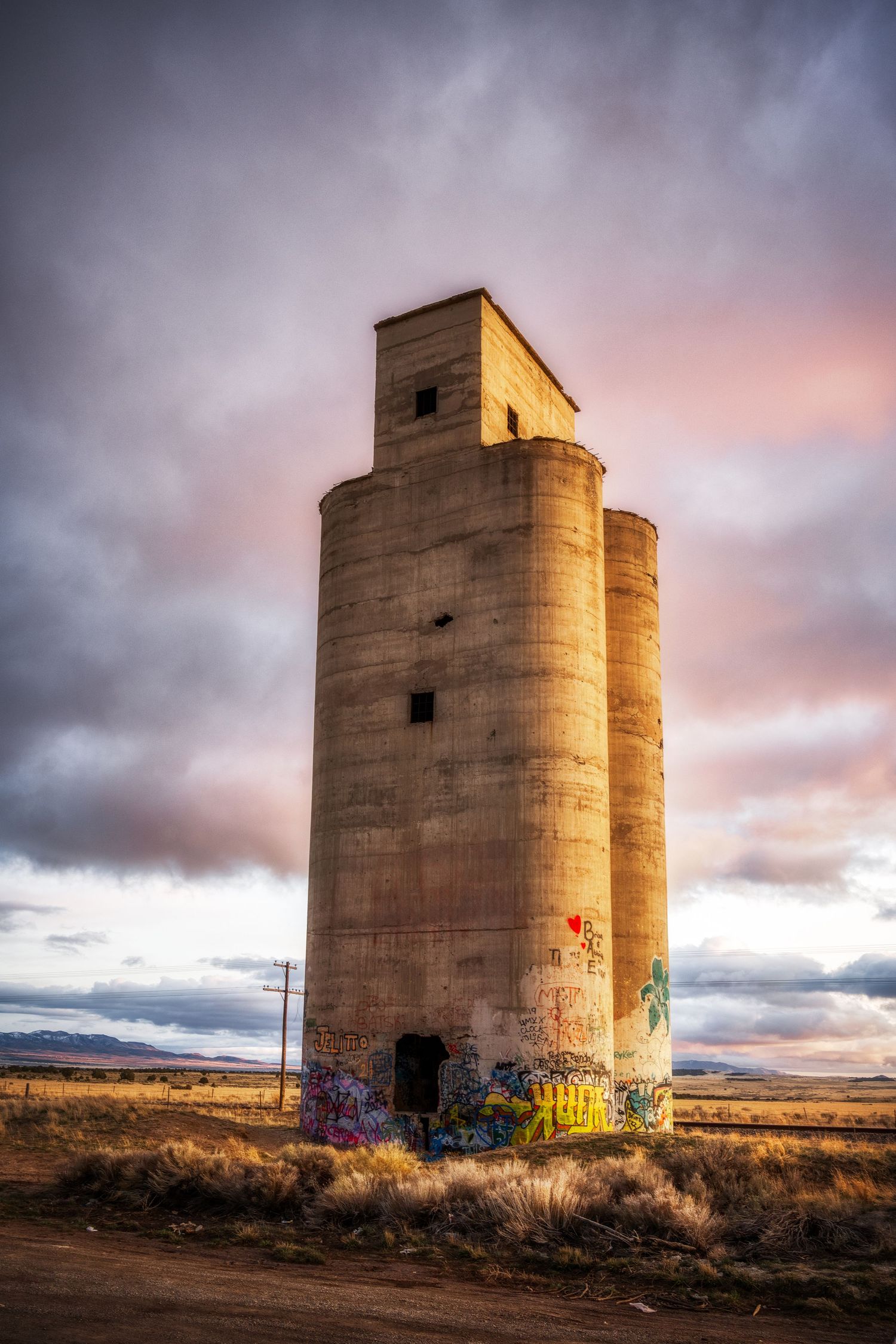 Abandoned grain silo standing tall against dramatic pink sunset sky in rural landscape.