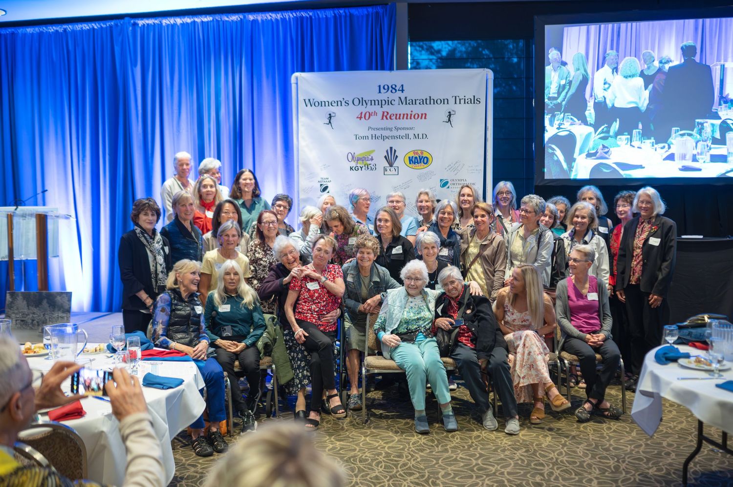 Large group photo of attendees gathered at a formal event with blue backdrop and presentation screen.