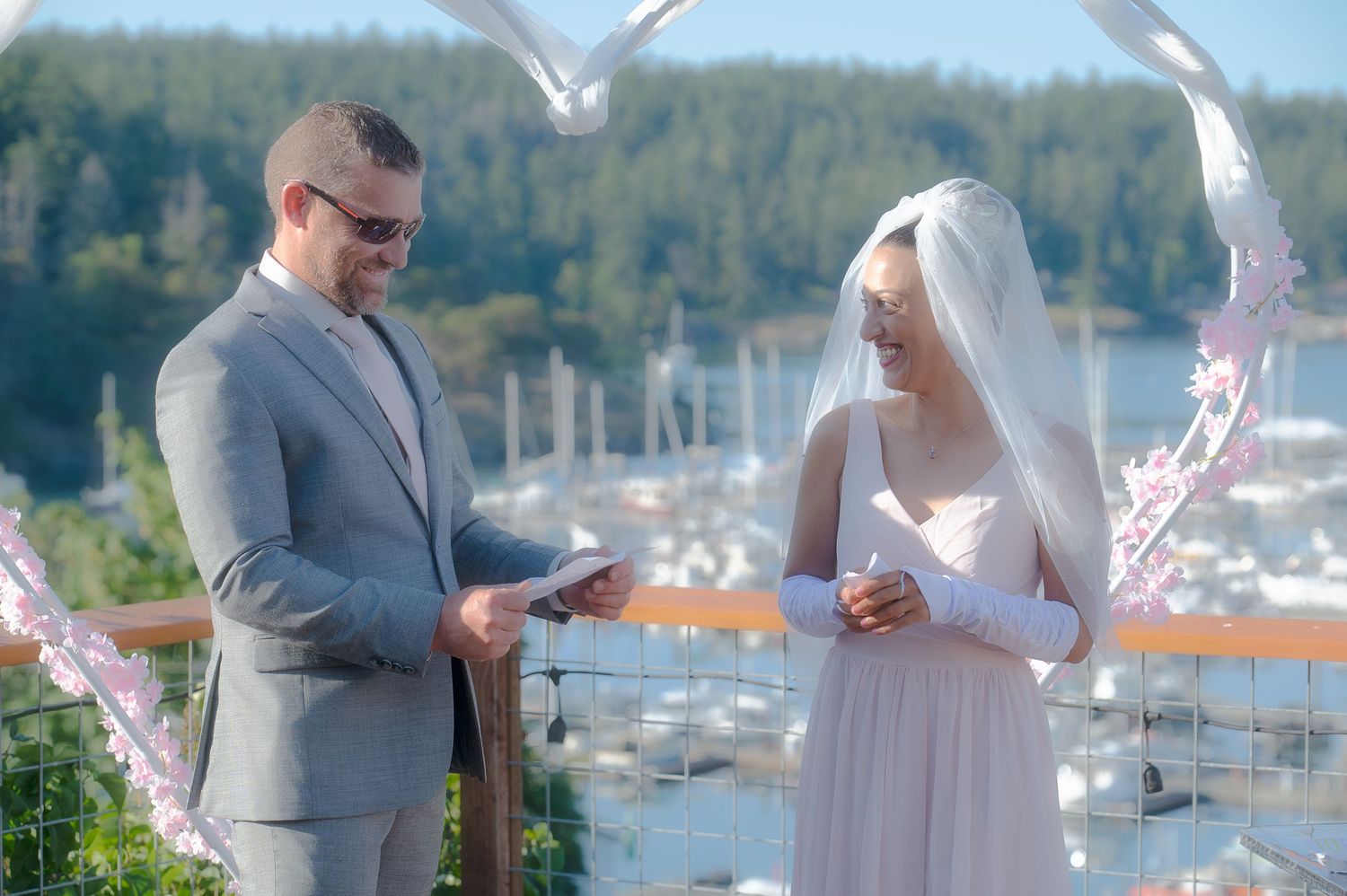 Couple exchanging rings at waterfront wedding ceremony with seagulls and marina in background.
