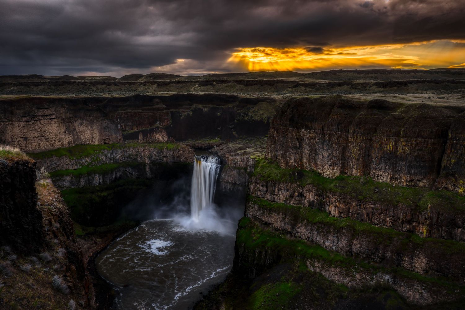 Majestic waterfall plunges down layered basalt cliffs at sunset with dramatic storm clouds in the background.