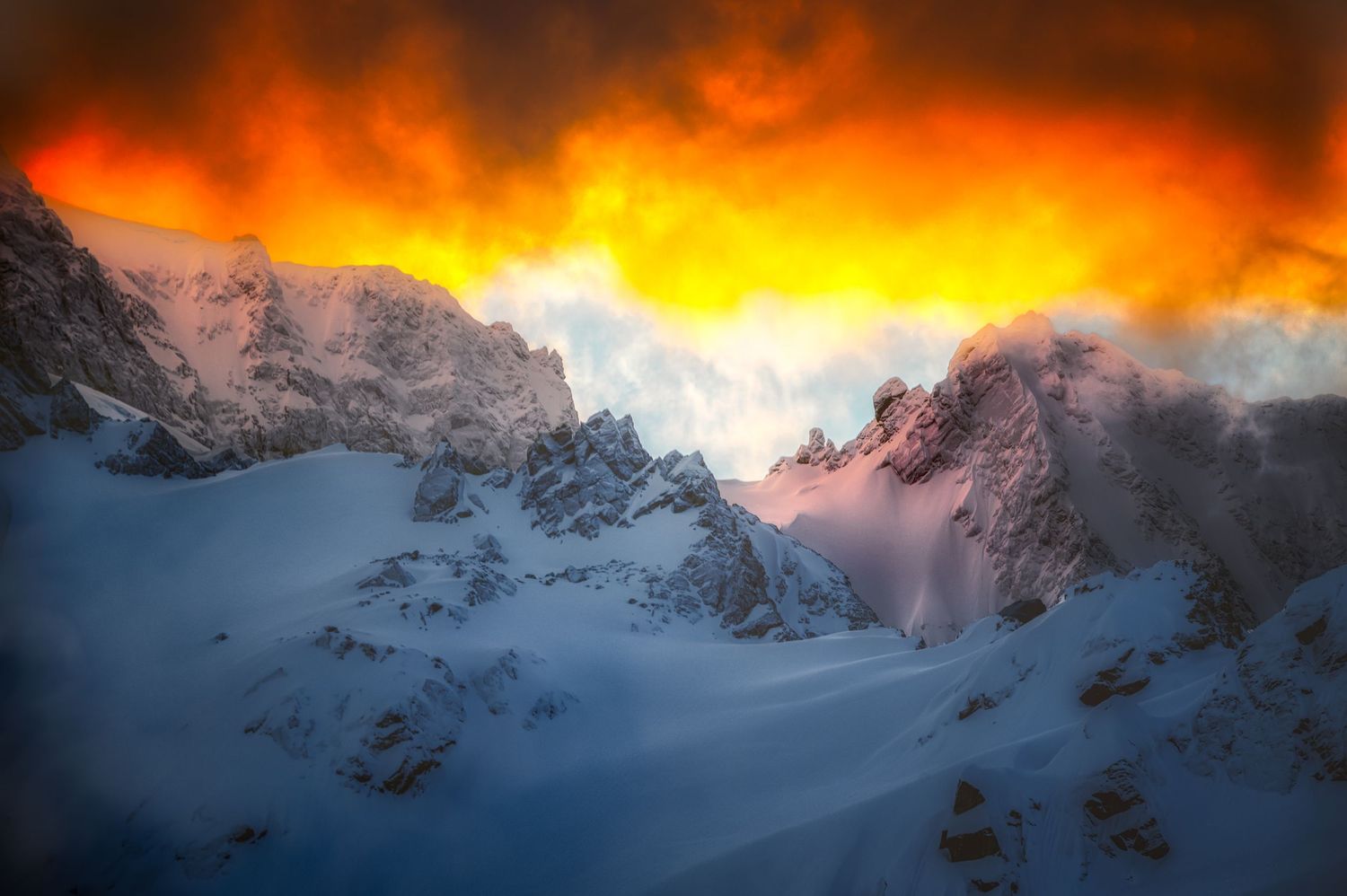 Fiery sunset illuminates snow-covered mountain peaks through dramatic orange and red storm clouds.