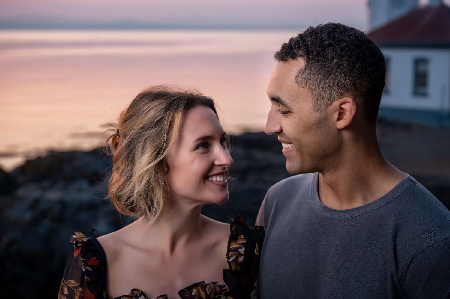 A romantic couple gazing at each other against a sunset coastal backdrop.