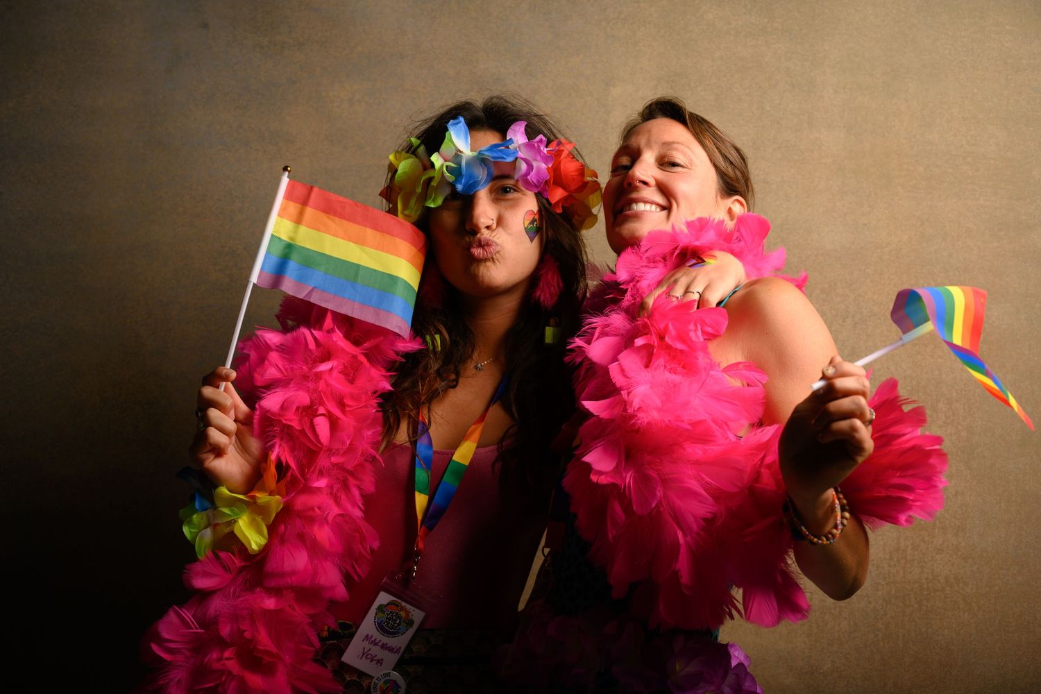 Two people celebrating with rainbow flags and pink feather boas against dark background.