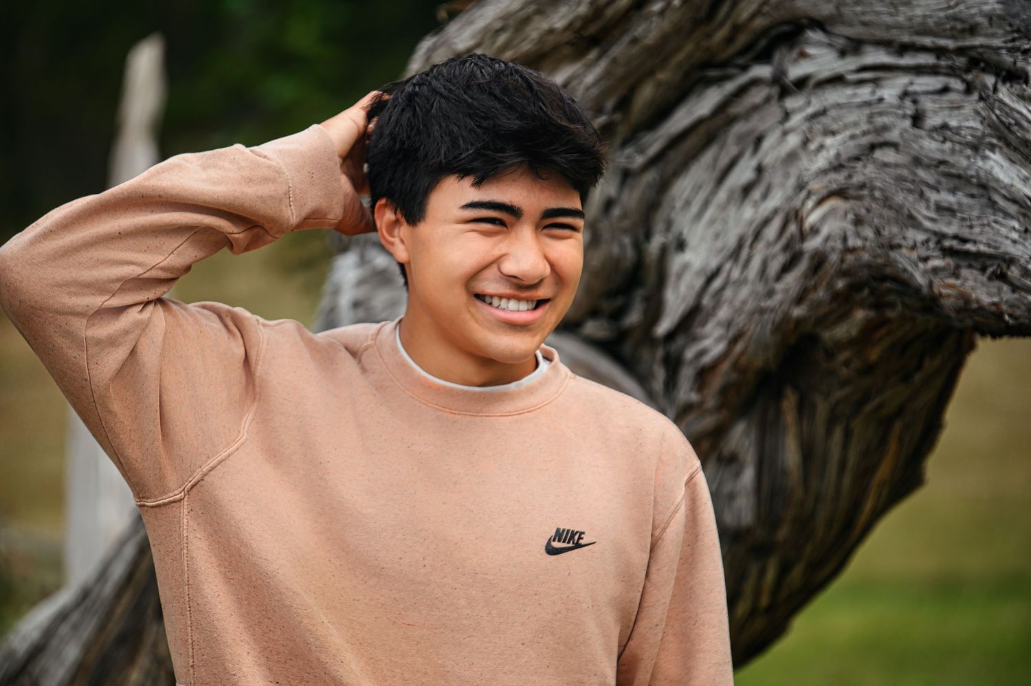 Person in beige Nike sweater smiling brightly while posing against a tree trunk.