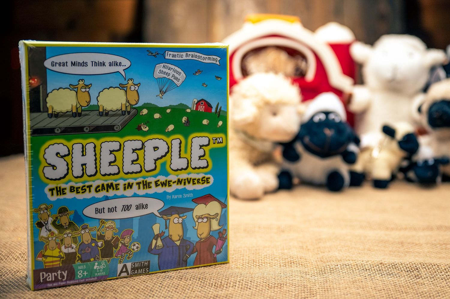 Sheeple board game box displayed with plush sheep toys in background on beige surface.