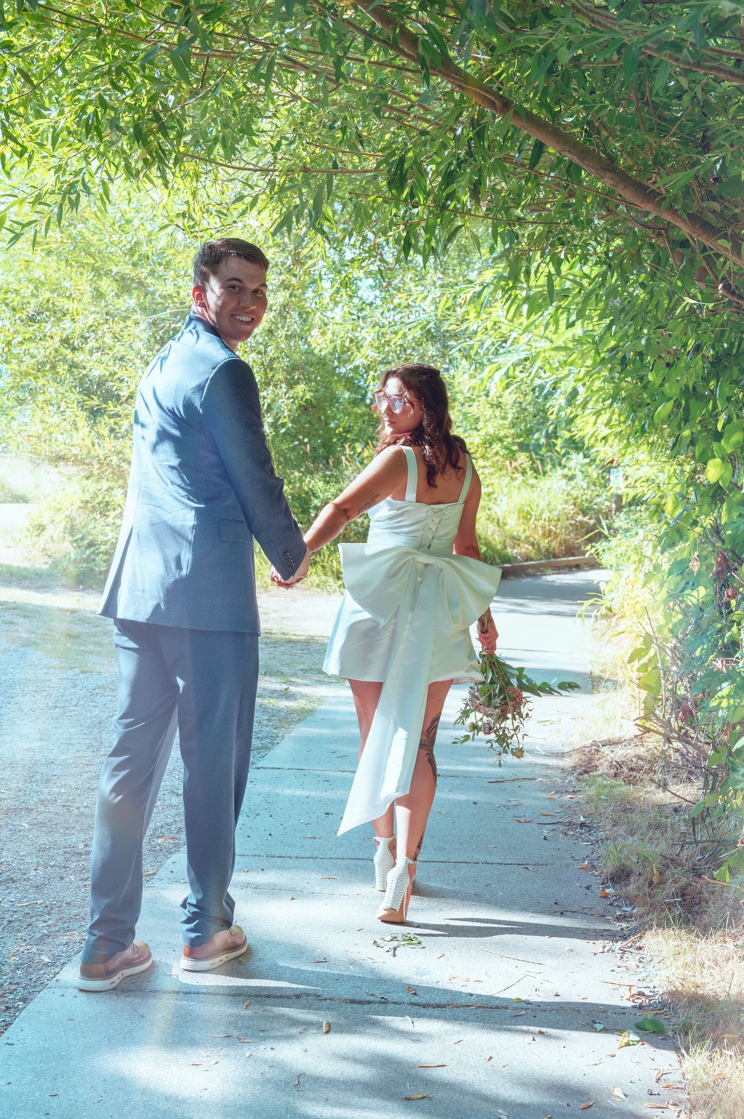 A couple enjoys a playful moment on a sunlit path under overhanging trees.