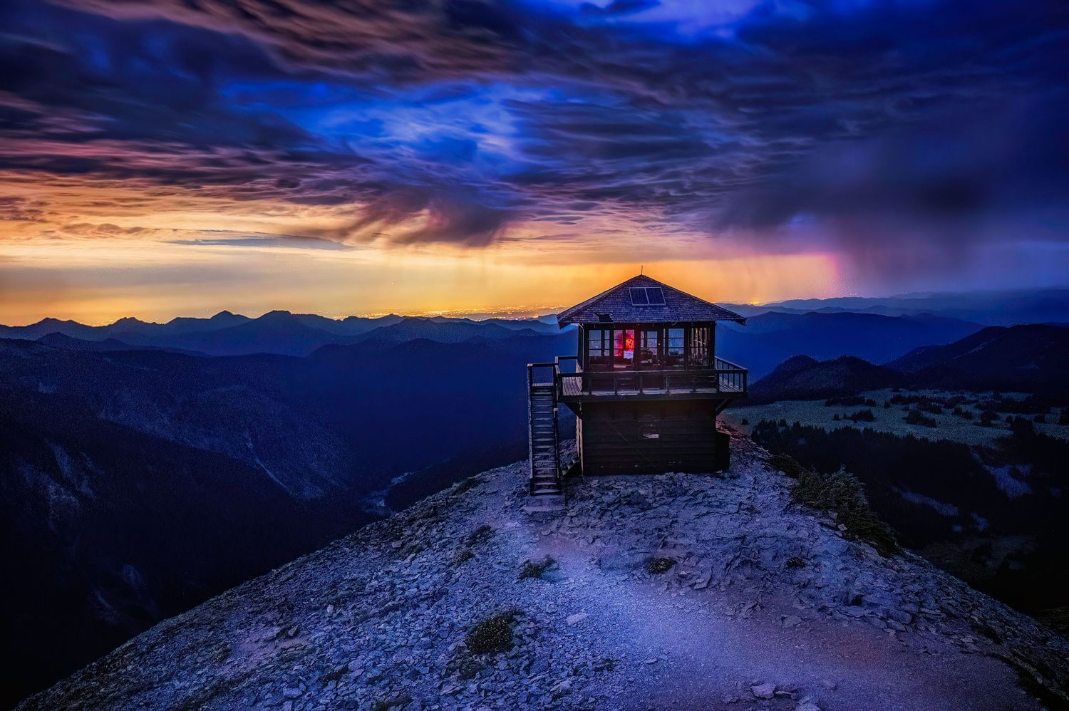 Fire lookout tower perched on mountain peak at sunset with dramatic storm clouds in the background.