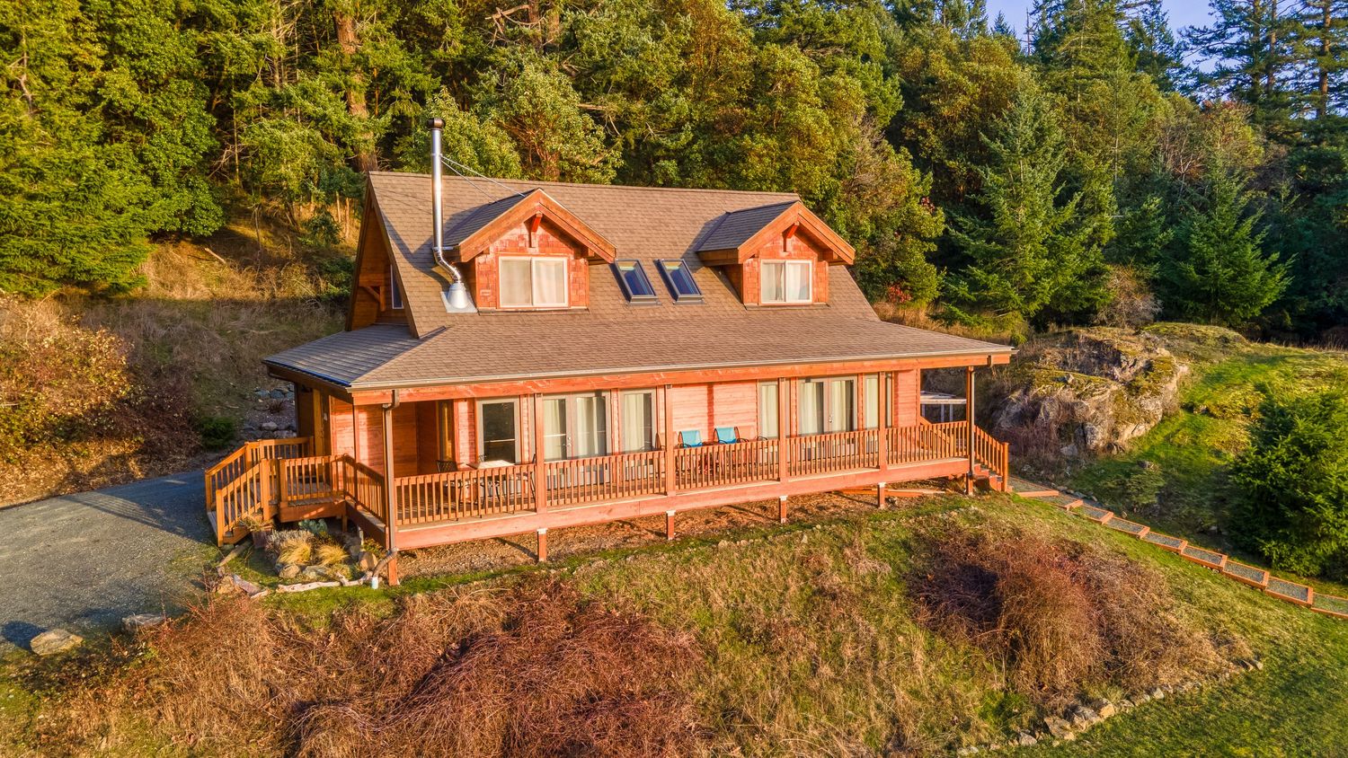 Rustic cedar cabin with wrap-around porch perched on a hillside surrounded by evergreen forest.