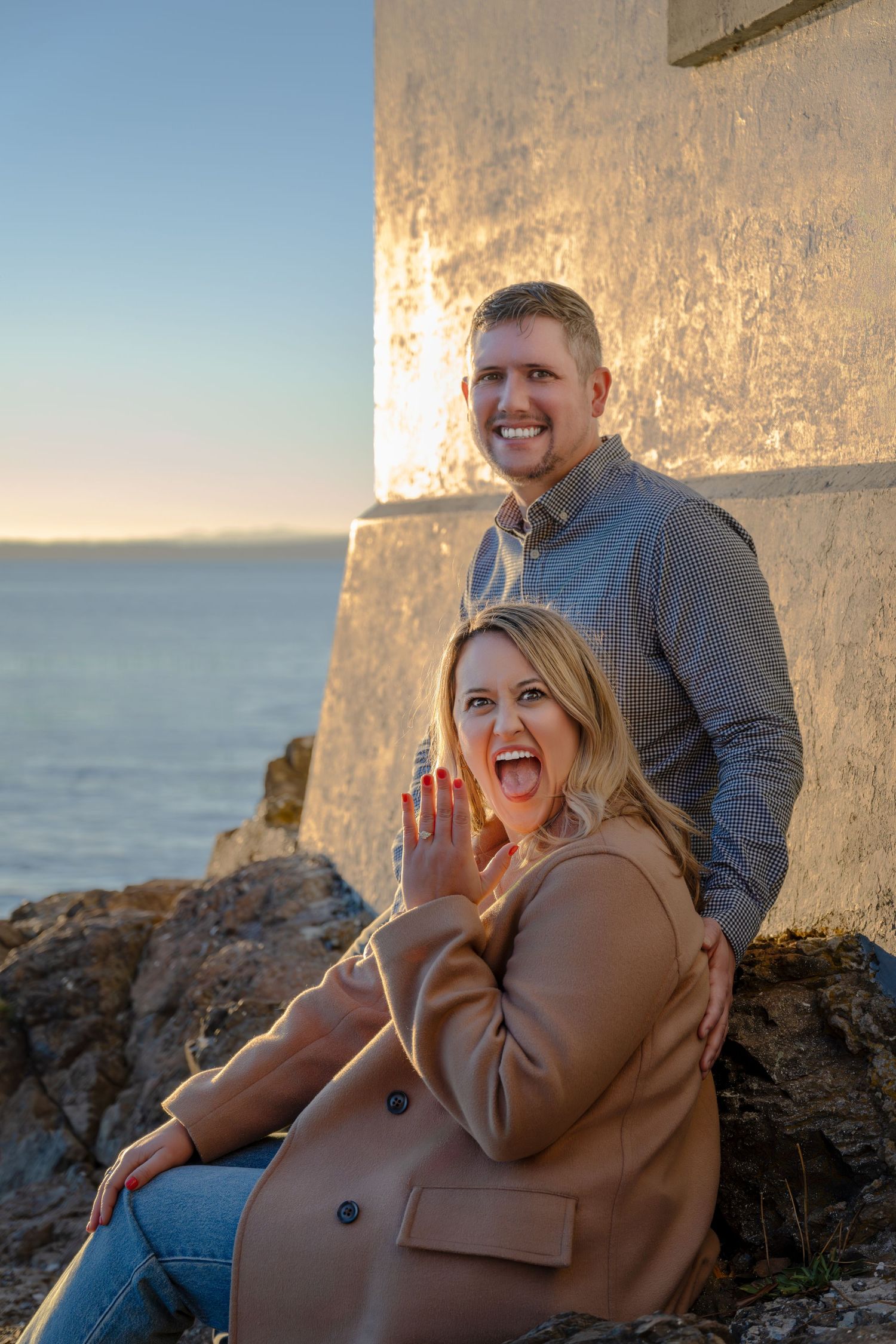 Happy couple poses together by lighthouse during golden hour.