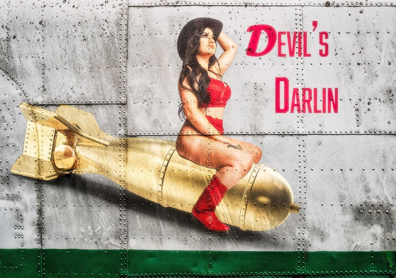 Pin-up style photo with figure in red sitting on golden bomb prop against industrial backdrop.