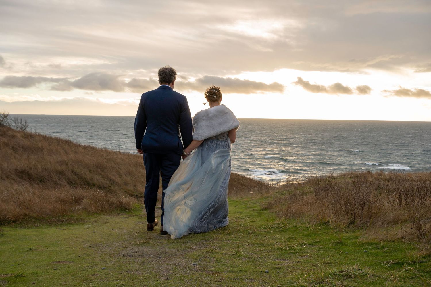 A couple walks along a coastal cliff path at sunset overlooking the ocean.