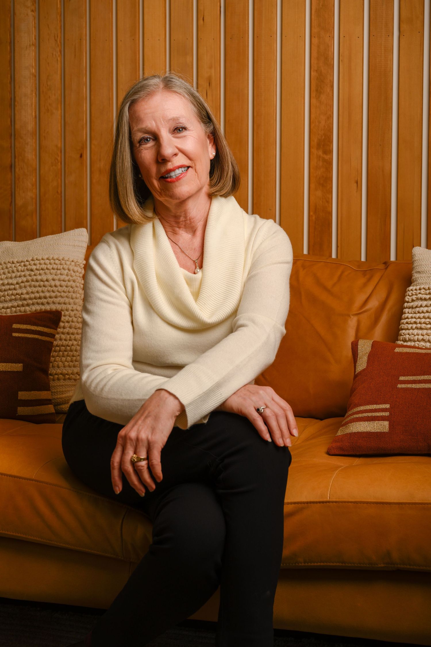 Person in cream cowl neck sweater sitting on tan leather couch with decorative pillows.