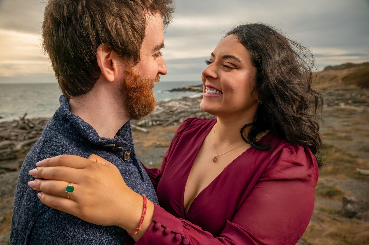Couple shares happy moment on coastal rocks, showing off engagement ring.