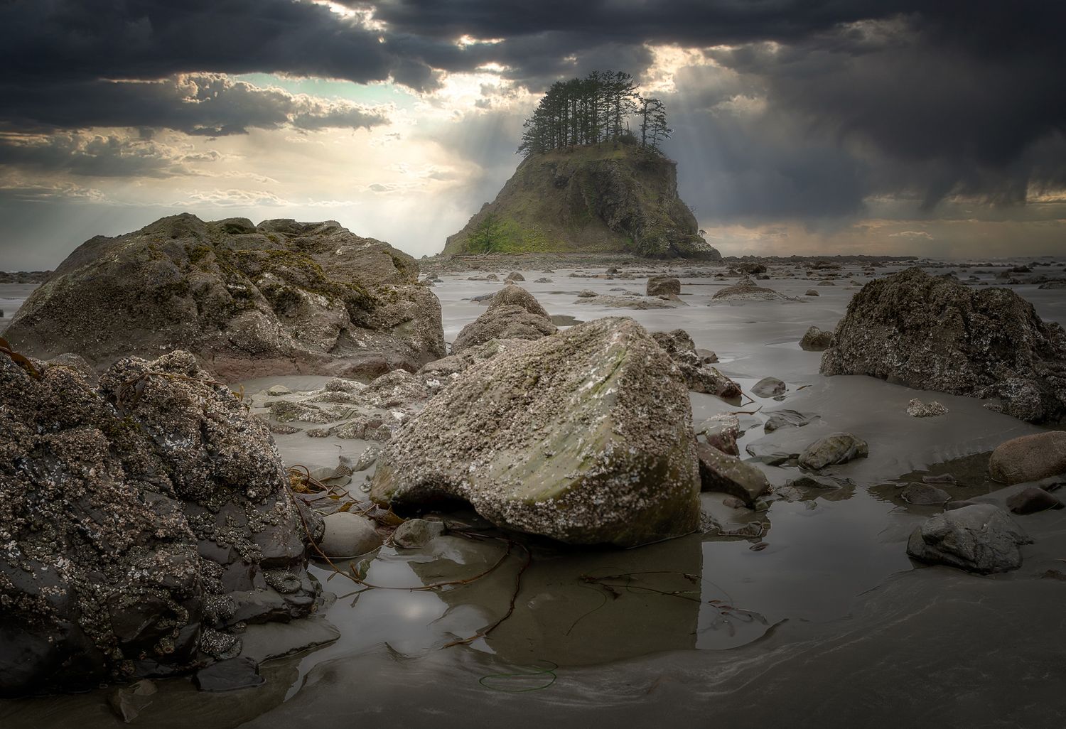 Dramatic sunbeams pierce storm clouds over a rocky island formation surrounded by turbulent waters.