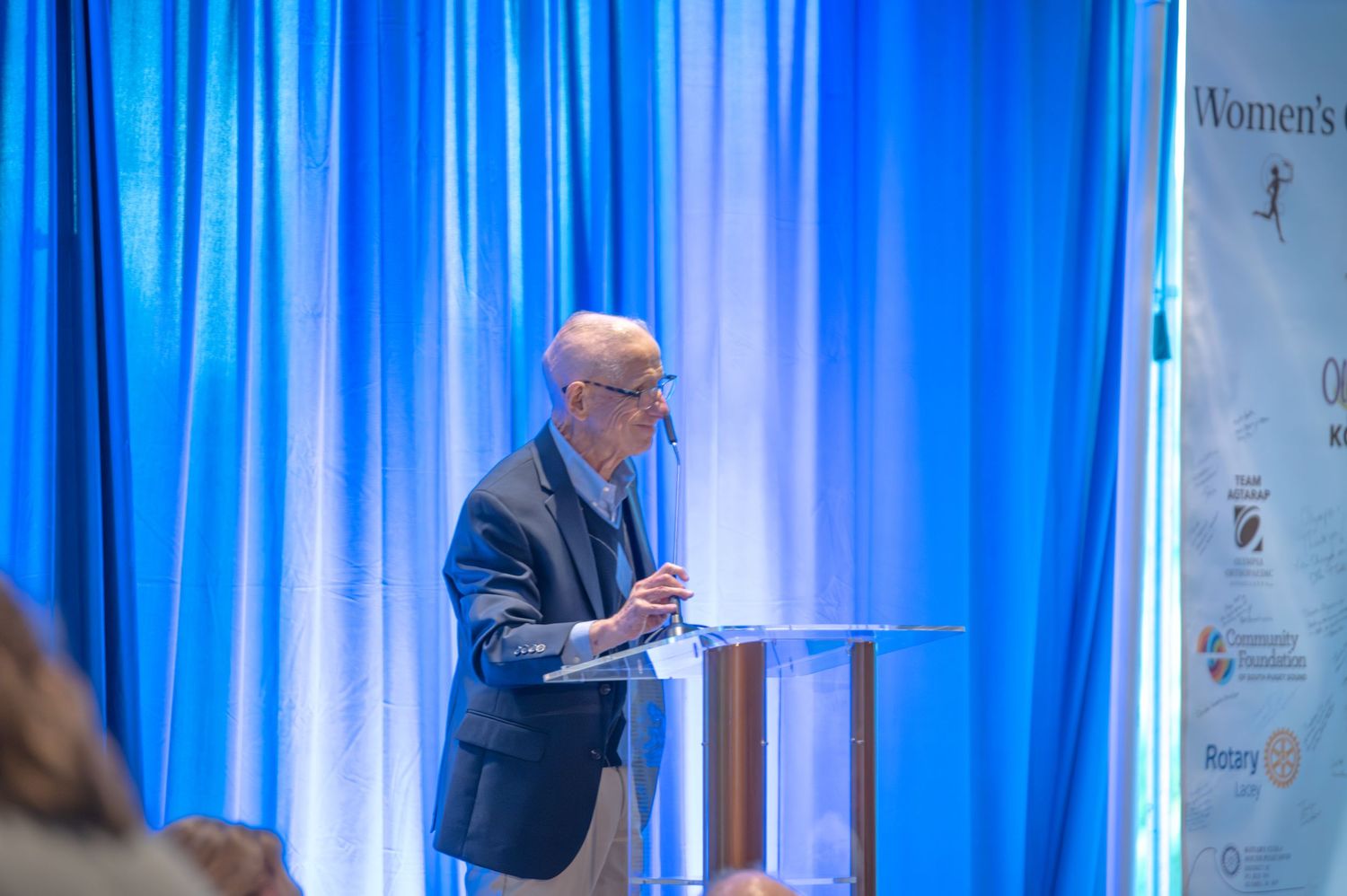 Speaker at podium presenting at event with blue curtain backdrop.