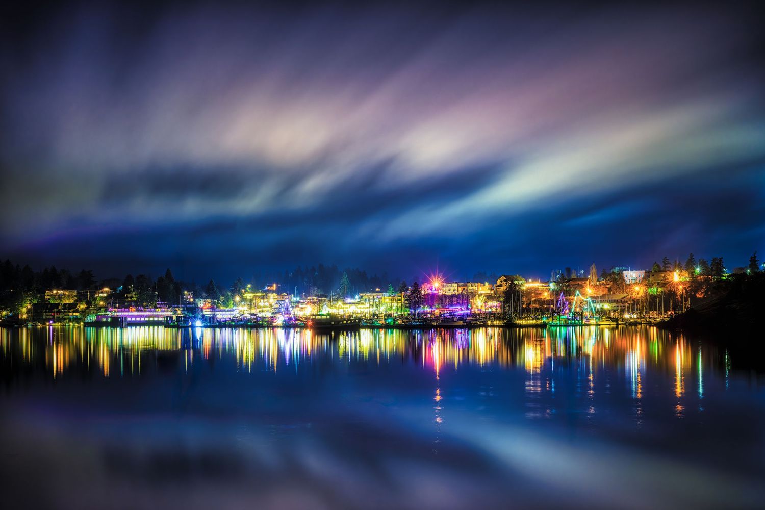 Colorful city lights reflect on calm water under aurora borealis in night sky.