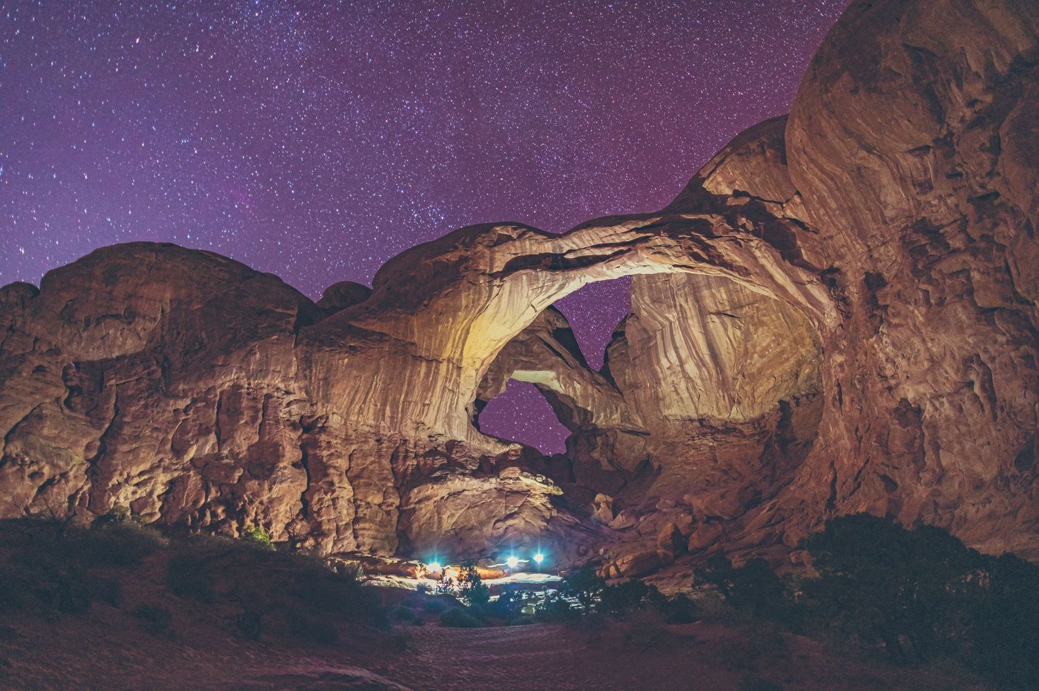 Natural rock arch formation silhouetted against starry purple night sky with glowing camp beneath.