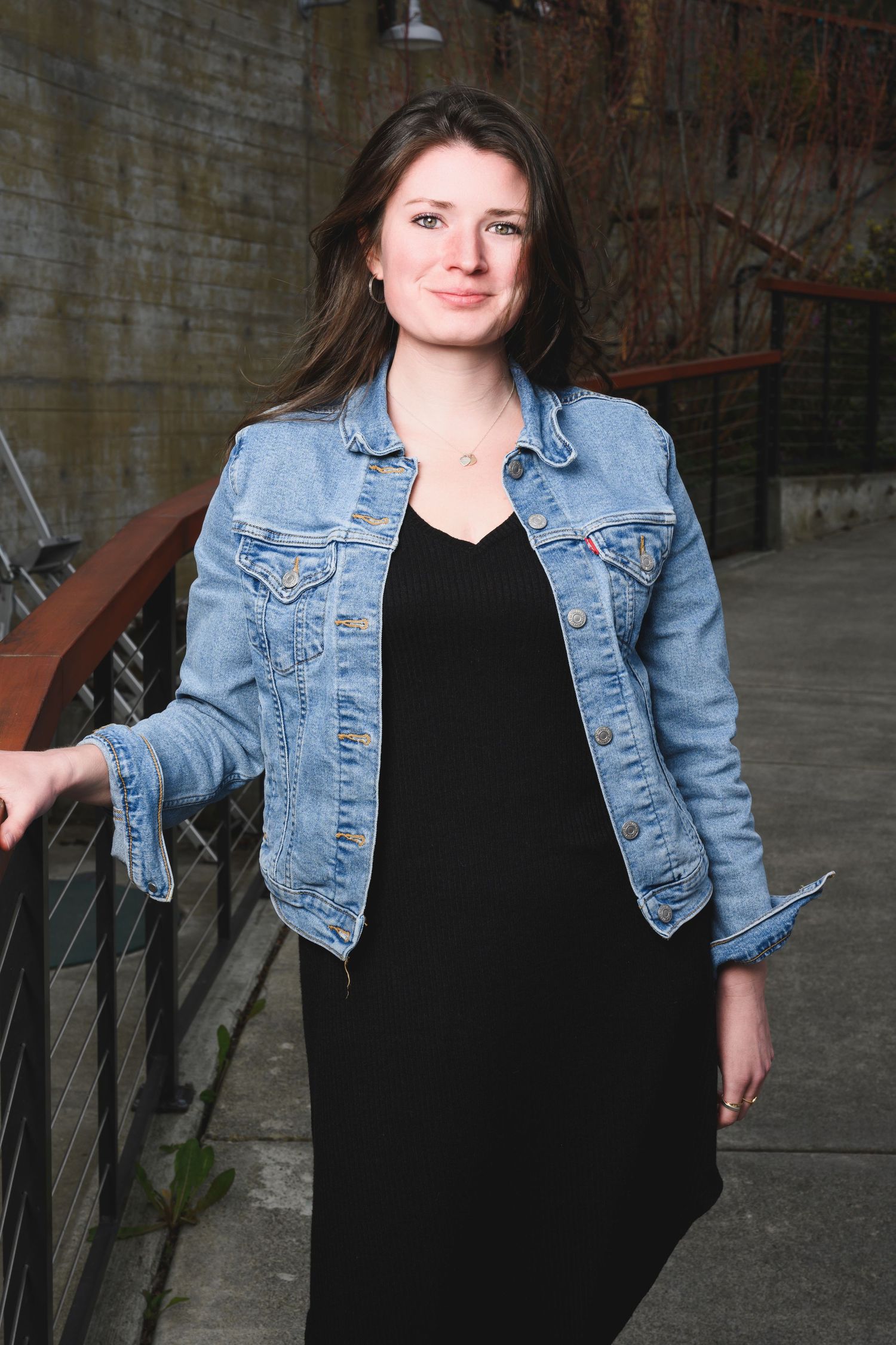 Person in denim jacket and black dress posing against industrial concrete wall background.