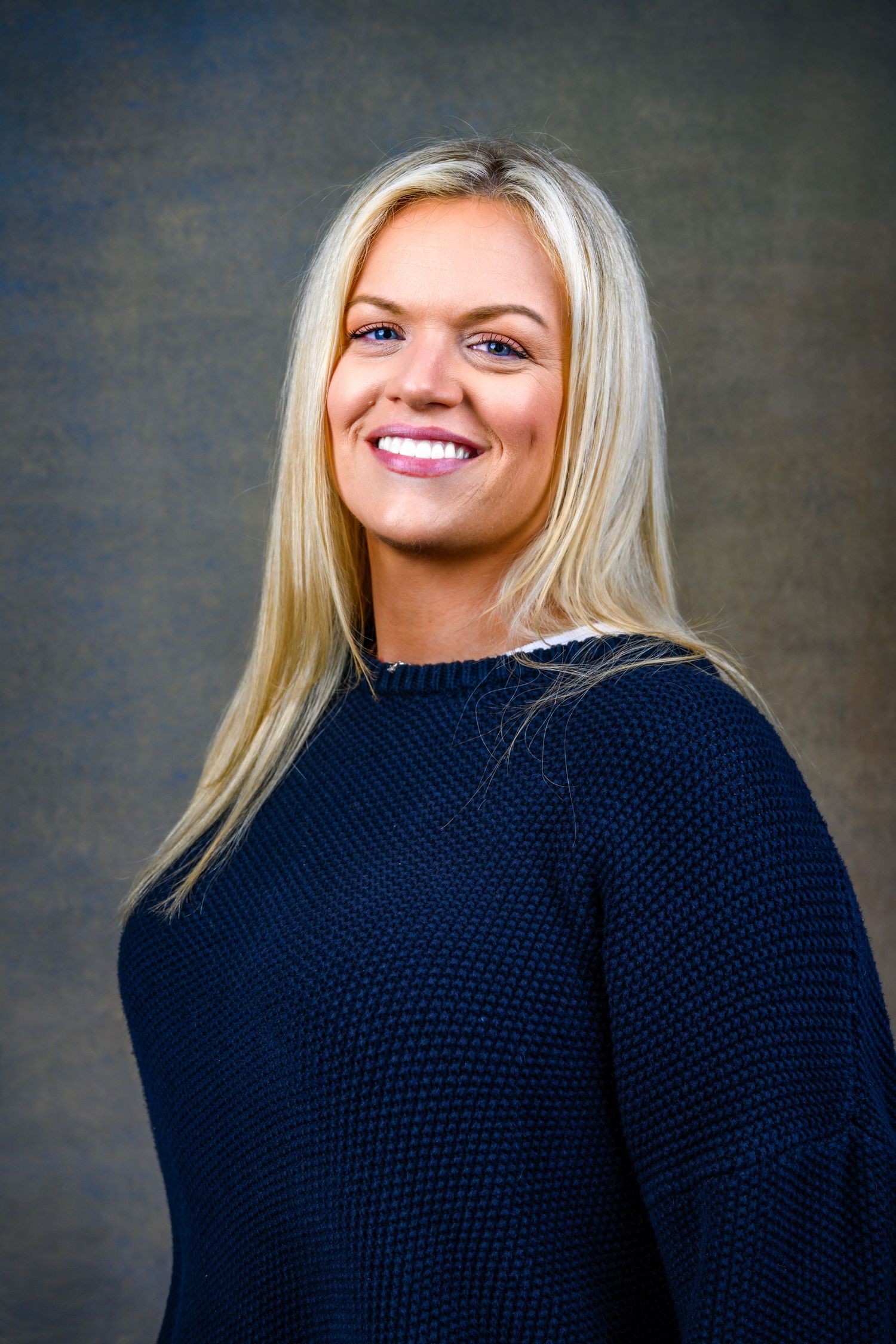 Professional portrait of person in dark knit sweater with long blonde hair and bright smile.