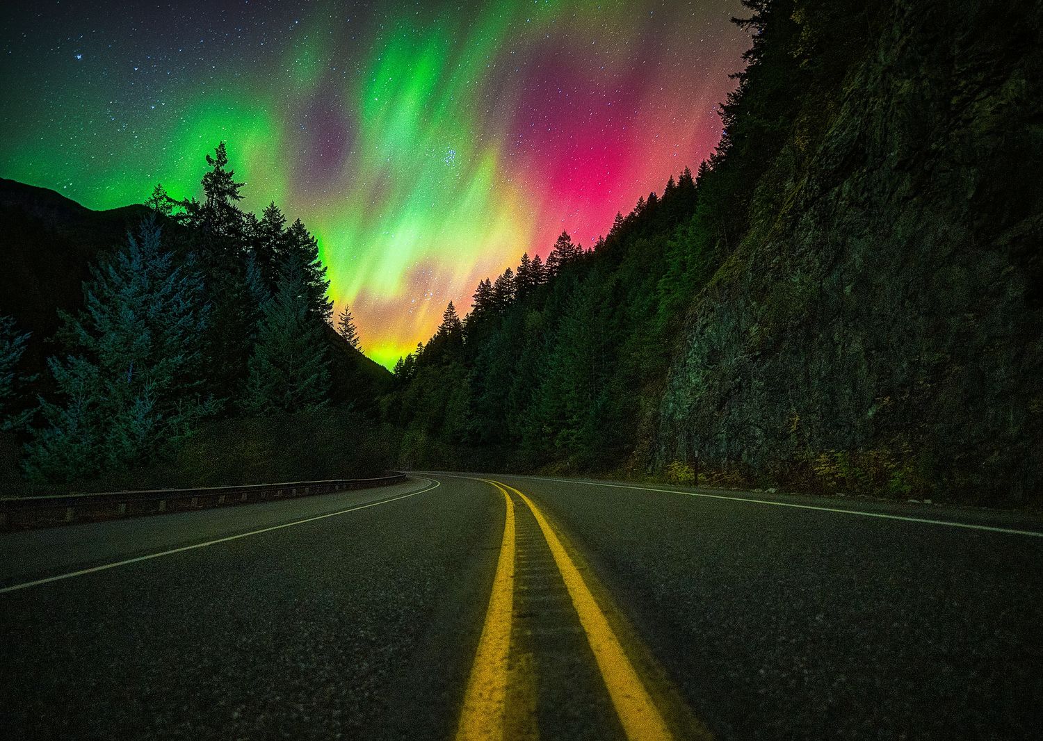 Aurora borealis casts green and pink lights over mountain highway through forest at night.
