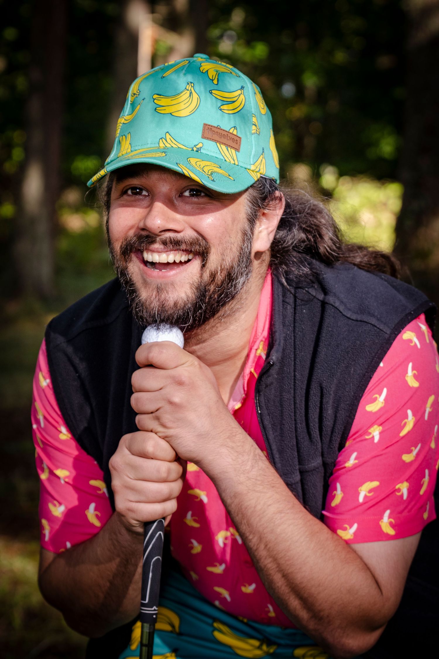 A performer wearing a playful banana-print hat and pink shirt holds a microphone while smiling outdoors.