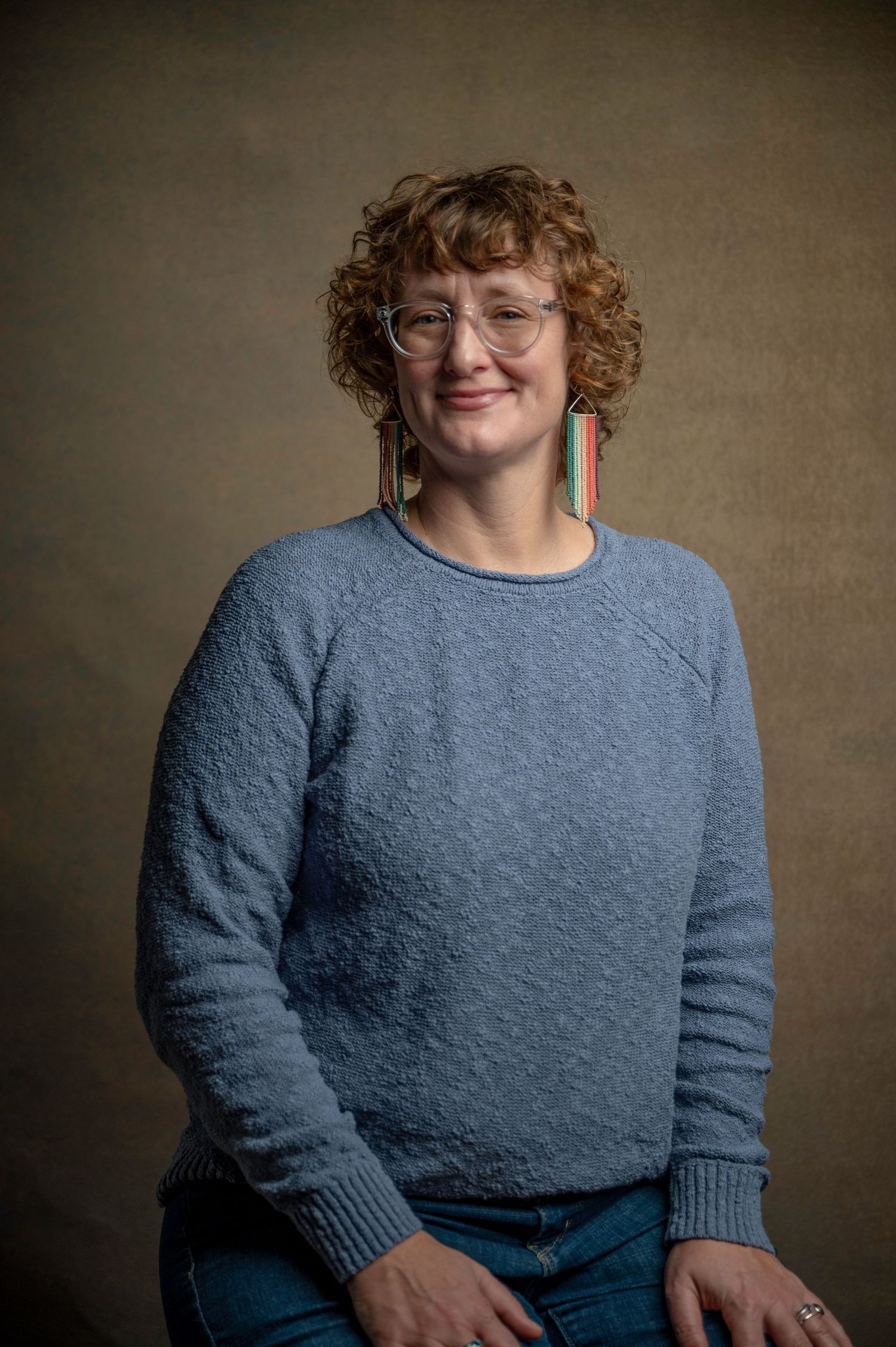 Portrait of person in blue sweater with curly hair and glasses against neutral backdrop.