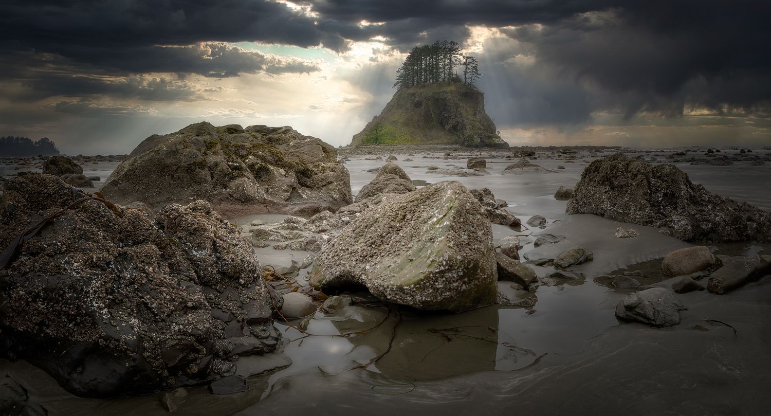 Dramatic seascape with sunbeams breaking through dark clouds over rocky coastal formations at low tide.