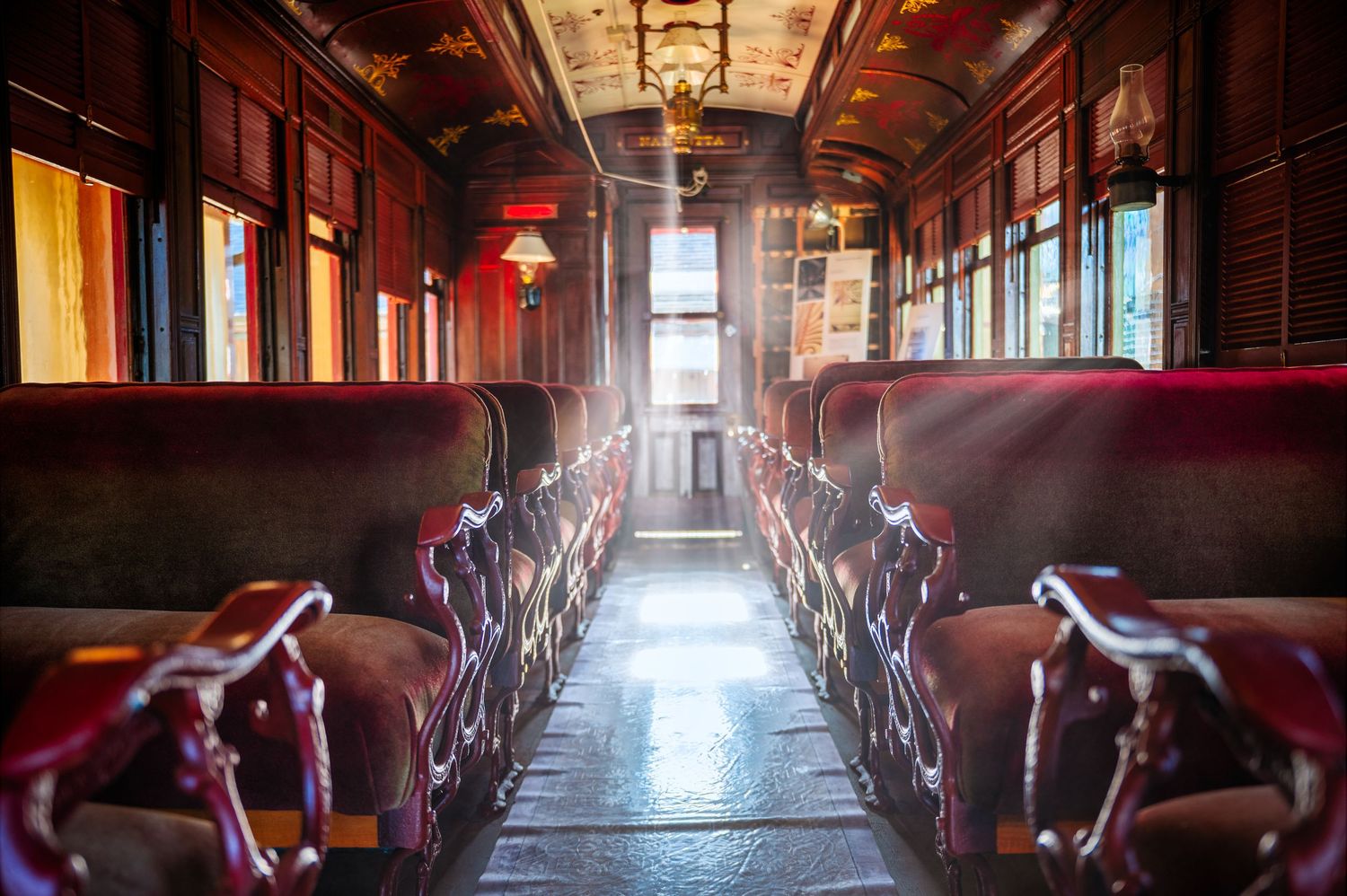 Vintage train car interior with red velvet seats and dramatic sunlight streaming through windows.