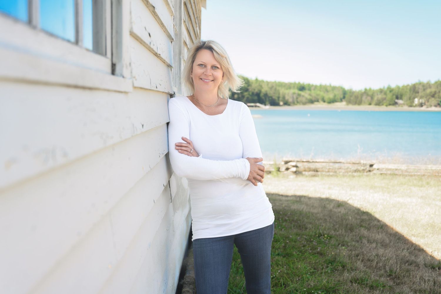 Person in white sweater leaning against white building with waterfront view.