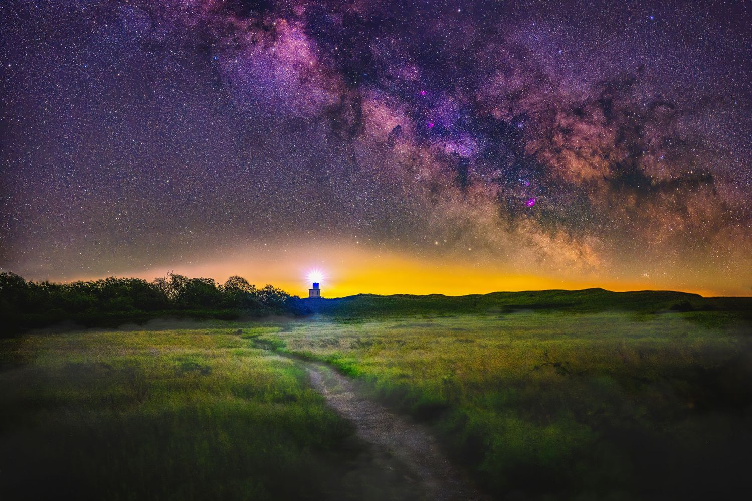 Vibrant Milky Way galaxy stretches across dark sky over illuminated path through grassy field.