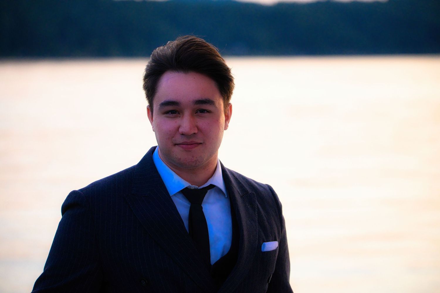 Professional portrait of someone in a dark suit and tie against a blurred waterfront background at sunset.