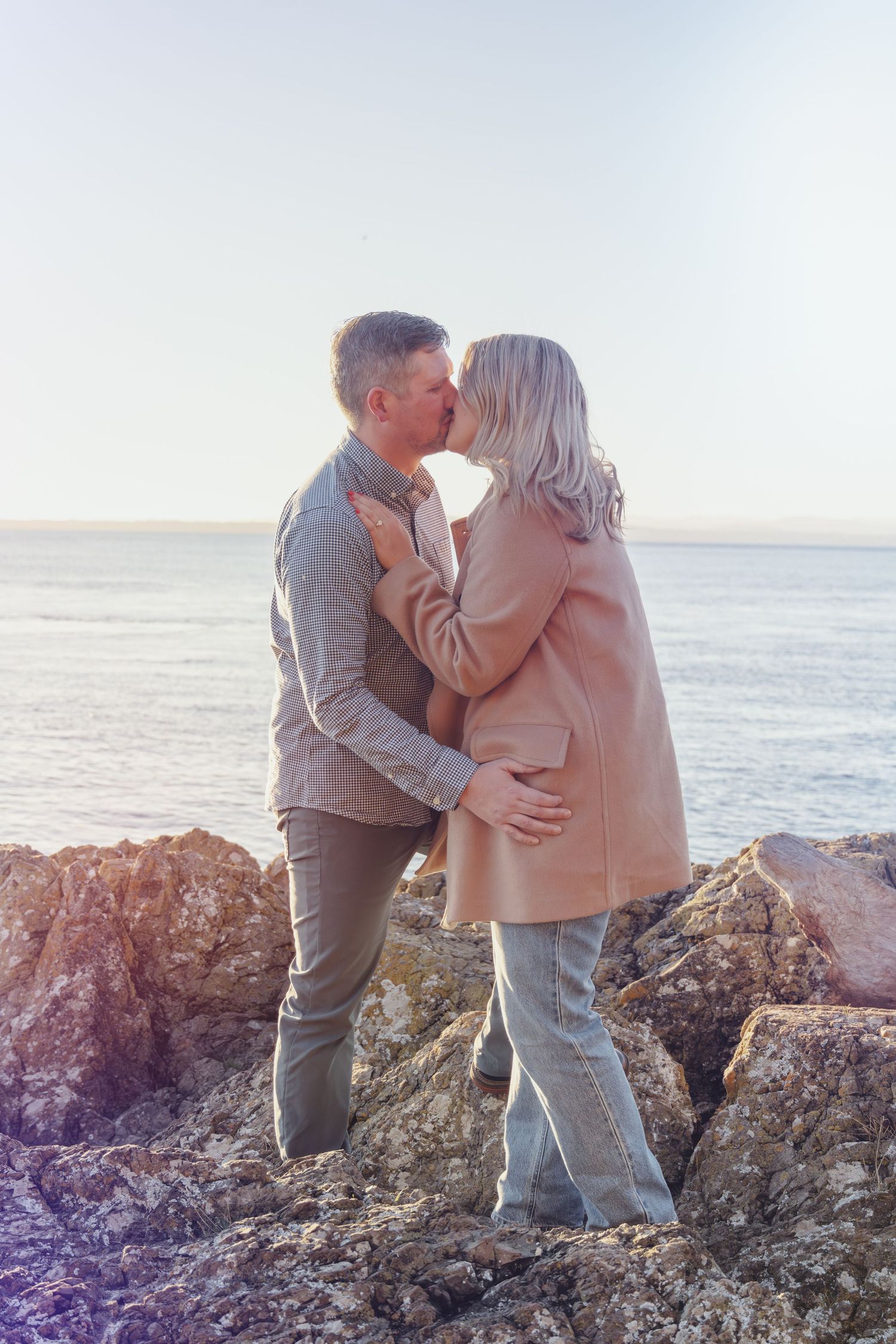 Romantic couple sharing tender moment on coastal rocks with ocean backdrop.