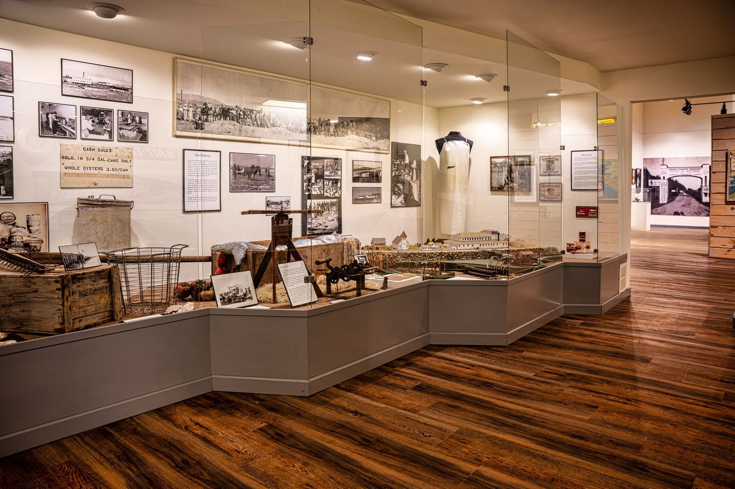 Historical museum exhibit displays artifacts and photographs with hardwood flooring and gray display cases.