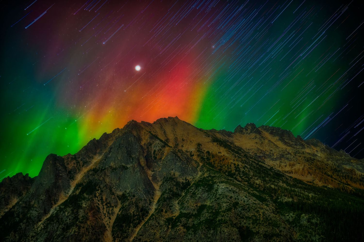 Star trails and aurora borealis create colorful light show over jagged mountain peak.