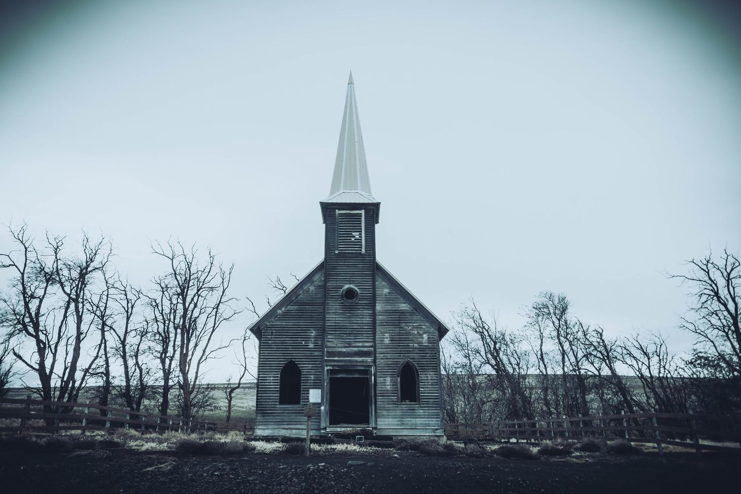 Abandoned wooden church with steeple stands alone in a desolate winter landscape under moody gray skies.