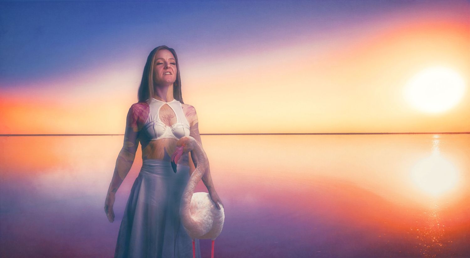 Silhouette in flowing dress holds glowing orb against serene sunset water backdrop.