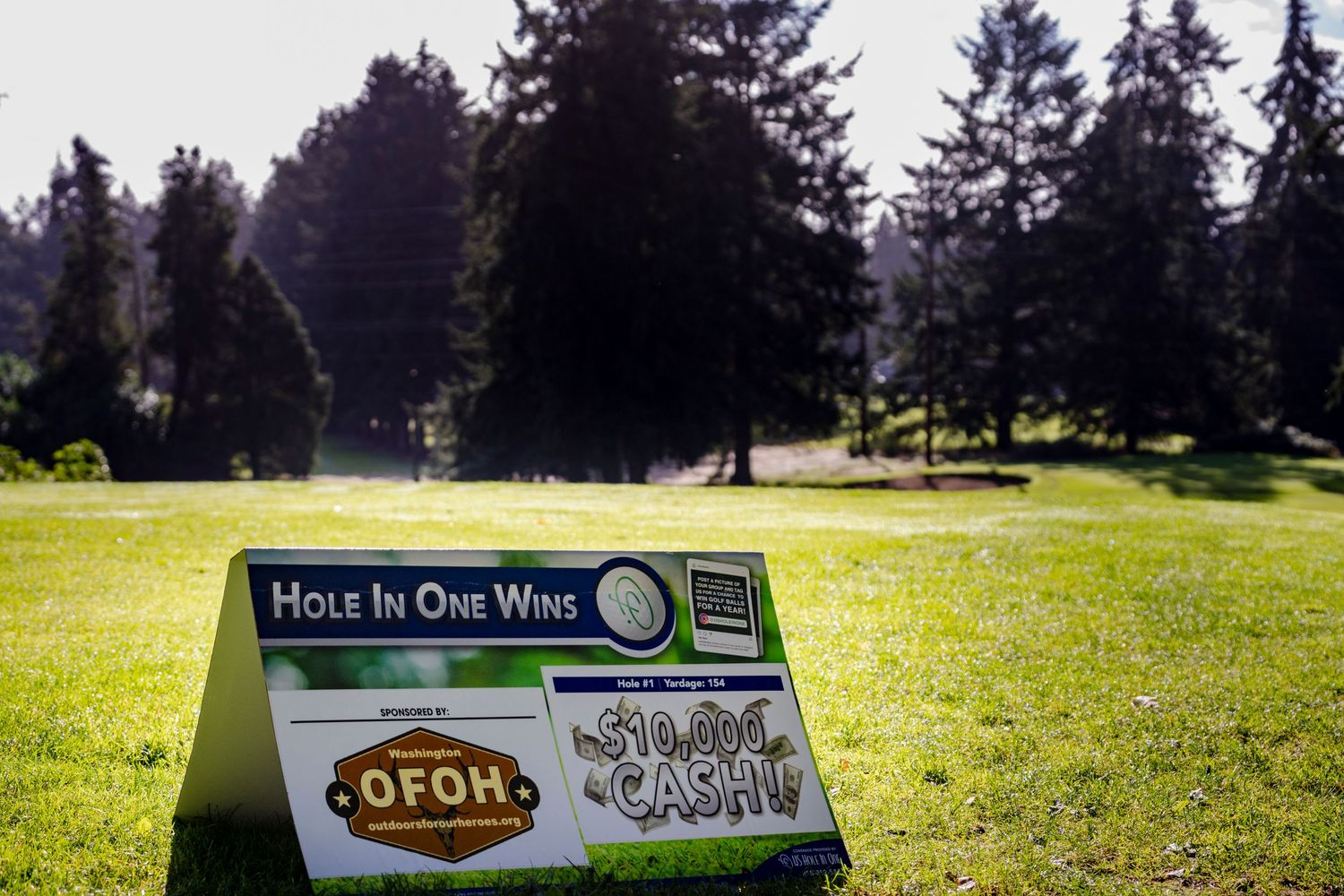 A hole-in-one contest sign displayed on a sunny golf course with evergreen trees in the background.