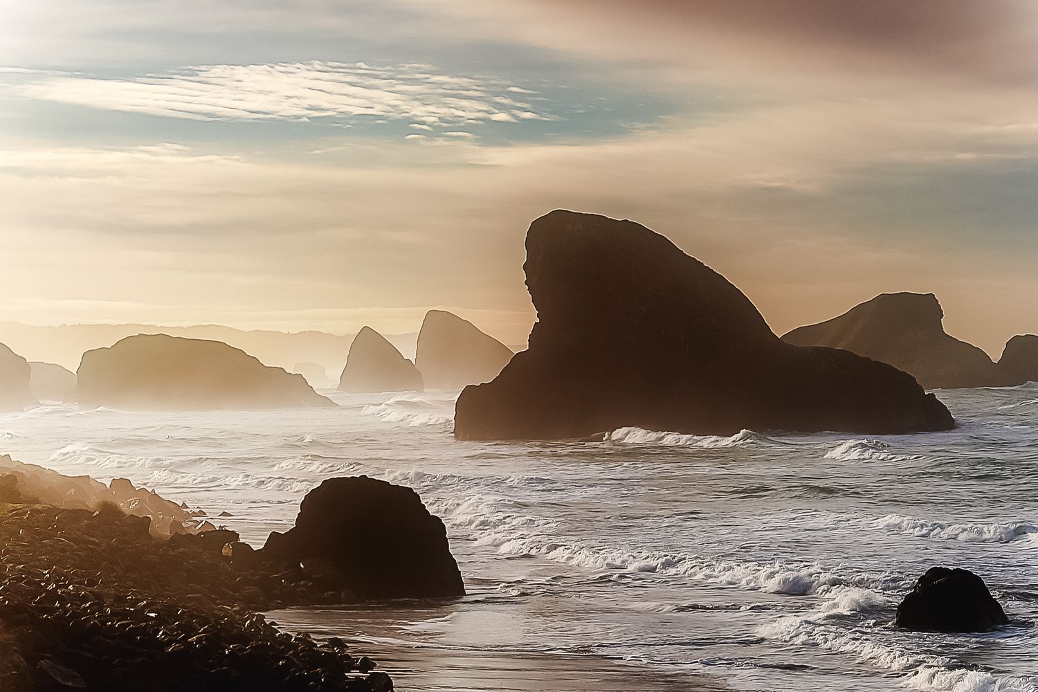Misty coastal seascape with large rock formations emerging from churning ocean waves at sunrise.
