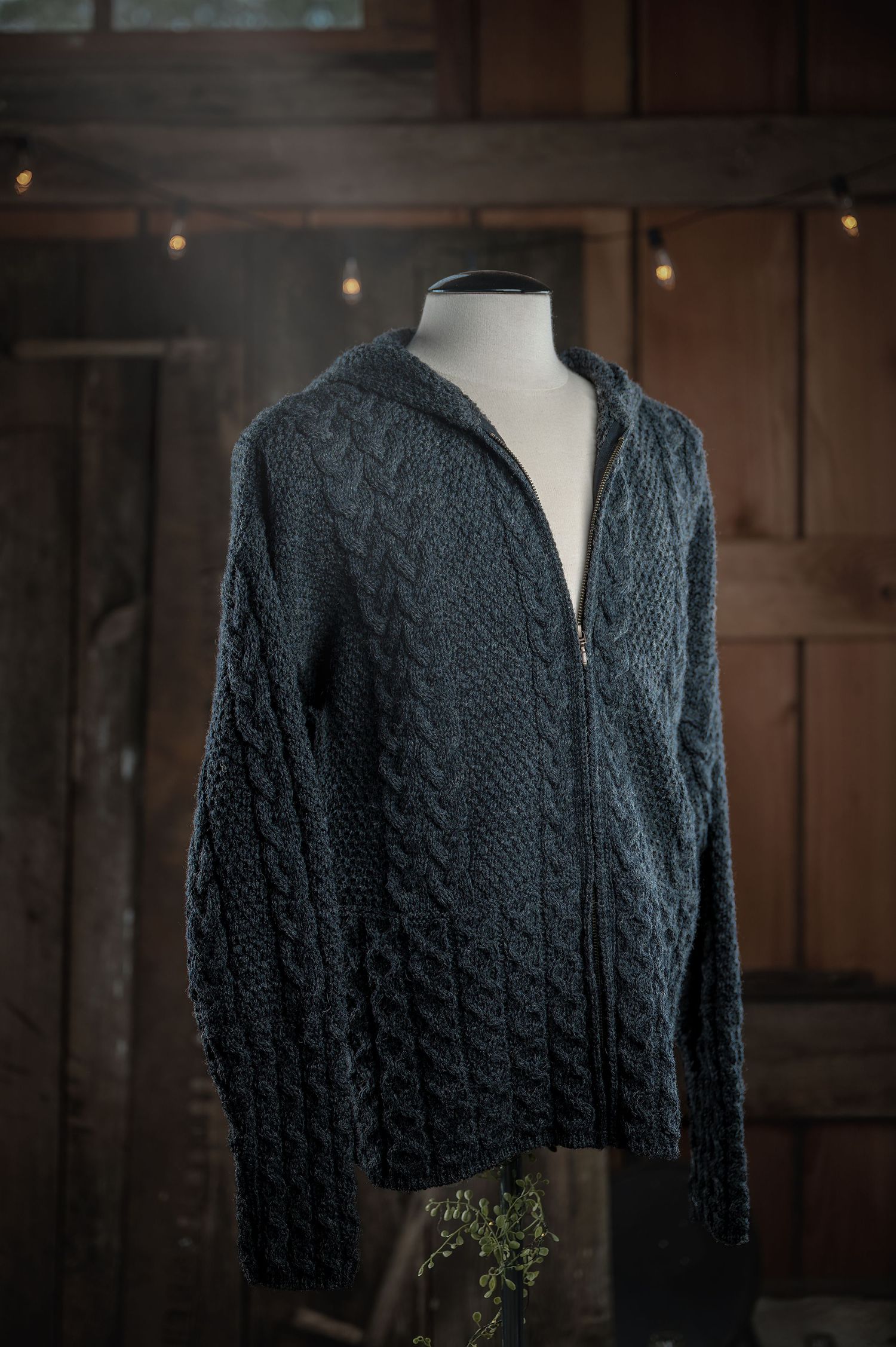 Gray cable knit sweater on mannequin form displayed against dark wooden wall with string lights.