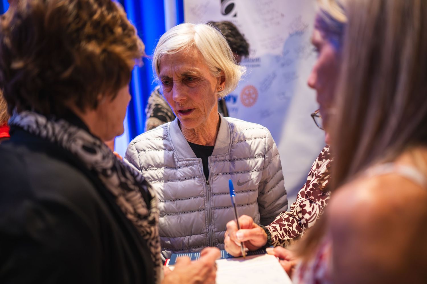 People engaged in conversation at an indoor event with blue curtain backdrop.