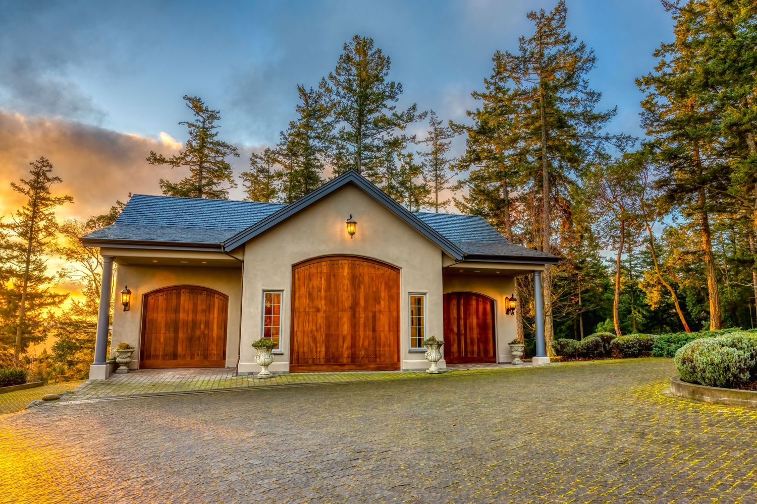 Charming stucco garage with dual wooden doors and dramatic sunset lighting through pine trees.