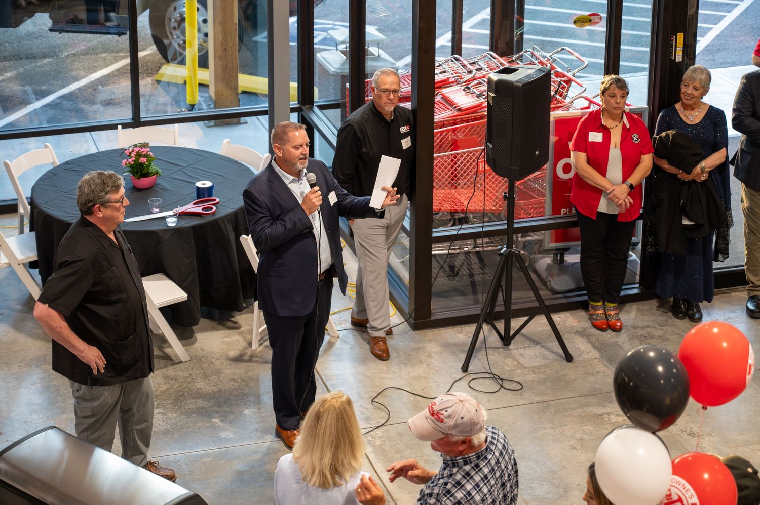 Group gathered around speaker at indoor event with red and black balloons for celebration.