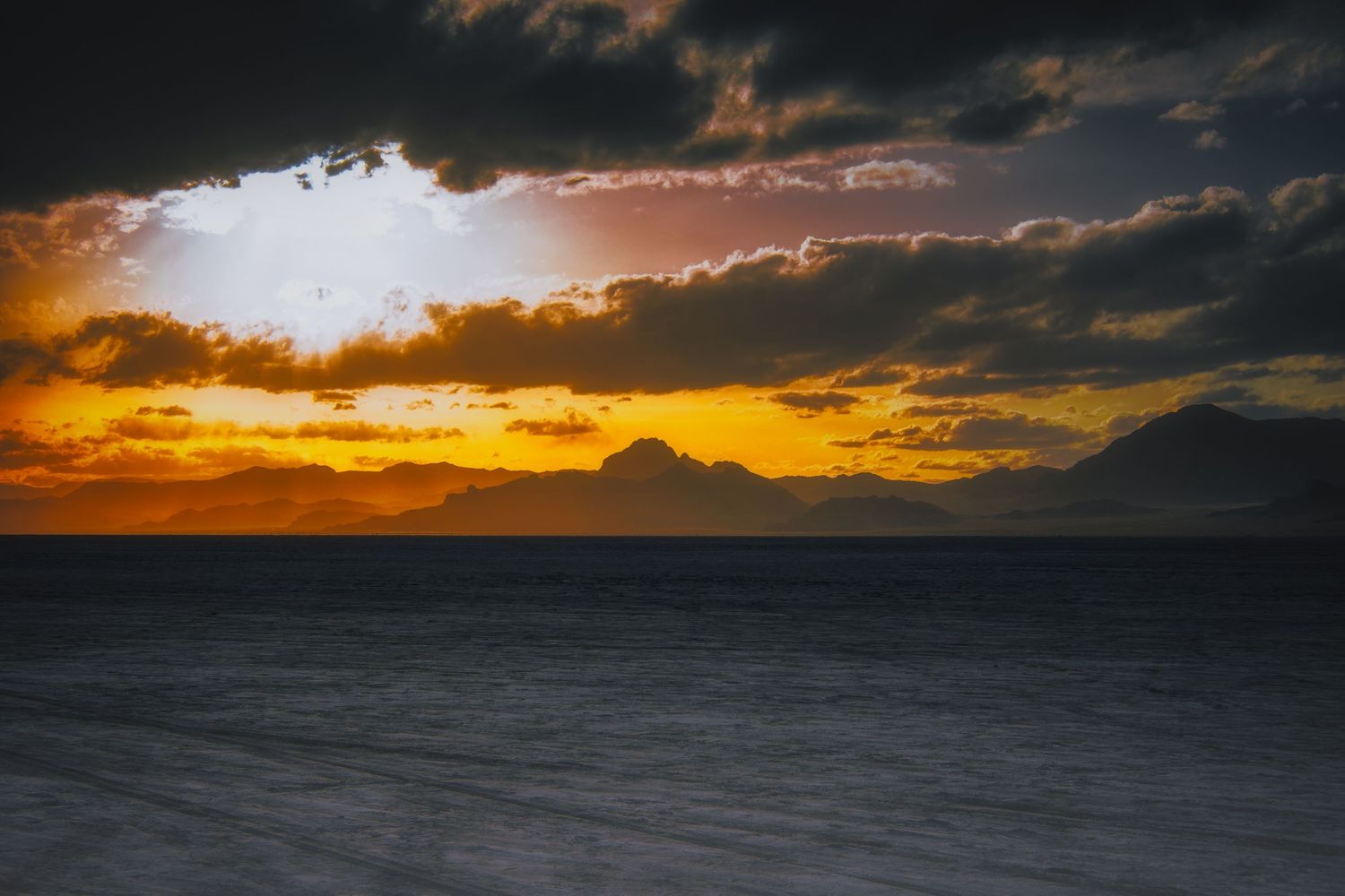 Vibrant orange sunset over dark ocean waters with dramatic cloud formations and mountain silhouettes.