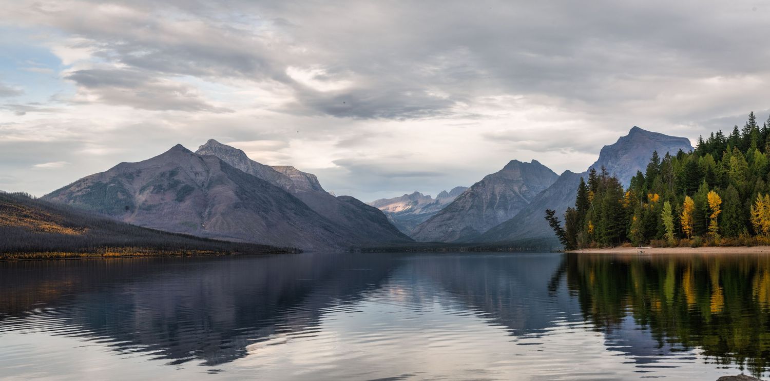 Perfectly still lake reflects snow-capped mountain peaks and forested shores under cloudy skies.